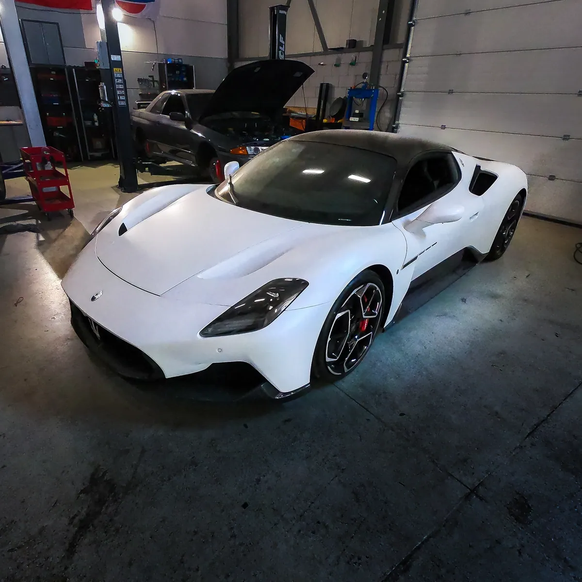 Witte supercar in de werkplaats van VPR-Engineering, met een auto op de brug op de achtergrond.
