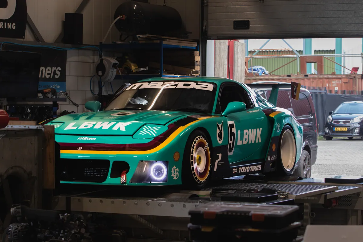 Turquoise Mazda RX-7 met Liberty Walk widebody, driekwart vooraanzicht in de werkplaats.