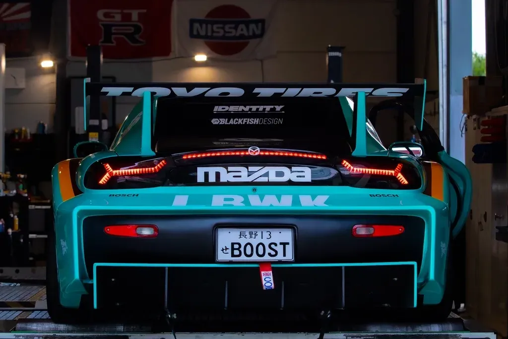 Achteraanzicht van de turquoise Mazda RX-7 met LBWK-vleugel en brandende achterlichten in de werkplaats.